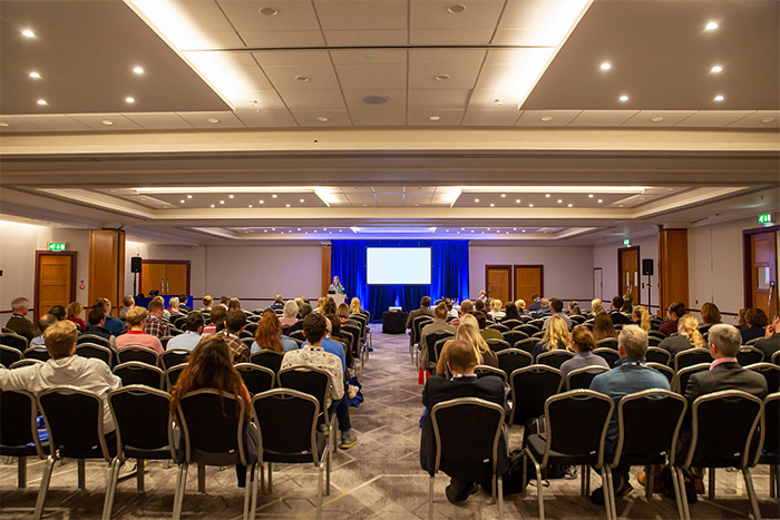 VPHA and AGV Conference audience seen from behind listening to speaker at lectern with large projector screen illuminated